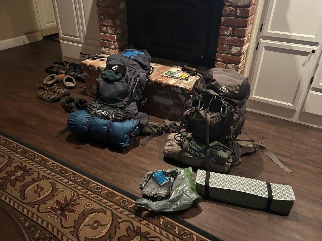 Two backpacks, sleeping bags, boots, and camping gear arranged on a living room floor during trip preparation
