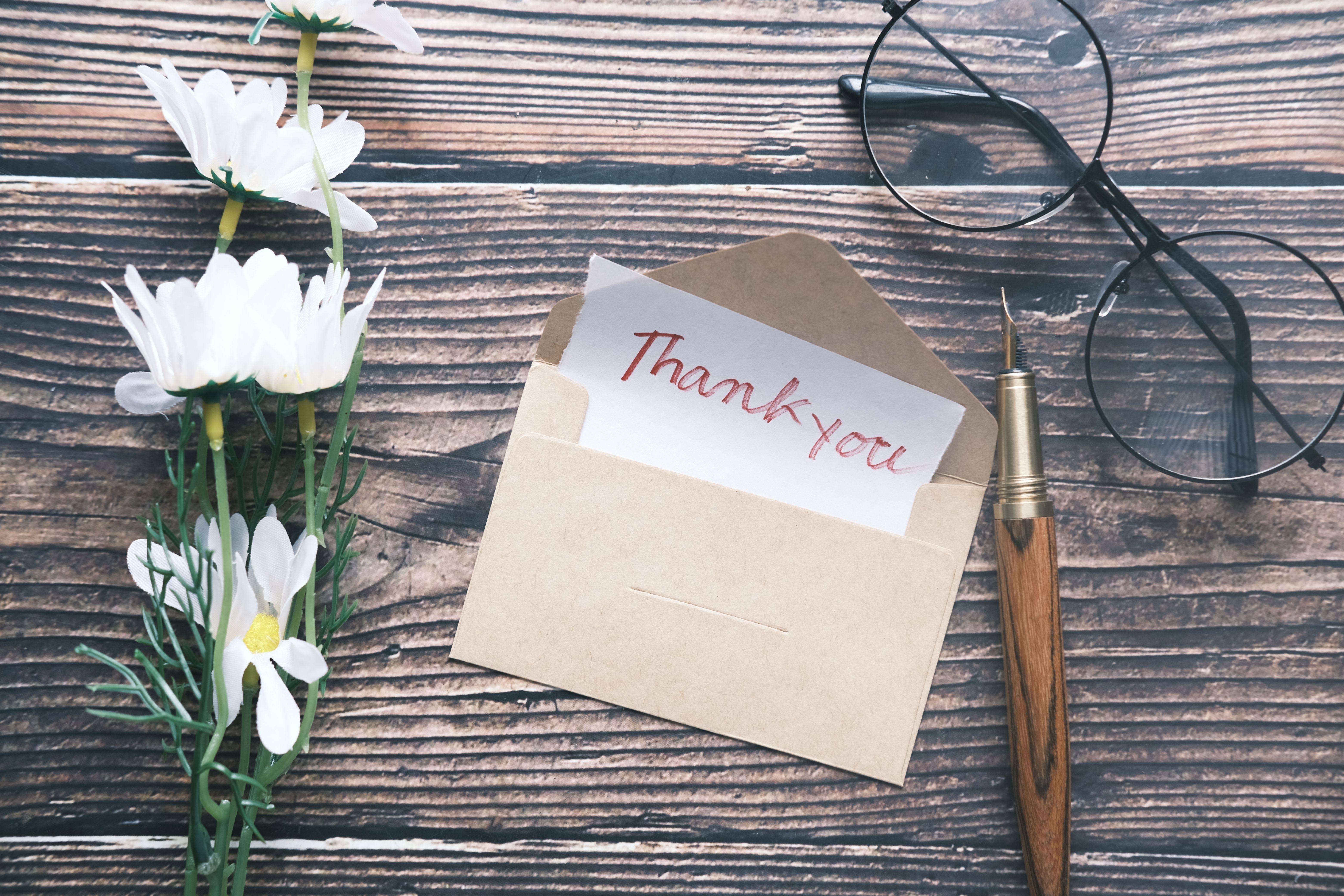 A handwritten thank-you note in a kraft envelope on a dark wooden table, beside white daisies, round glasses and a fountain pen.