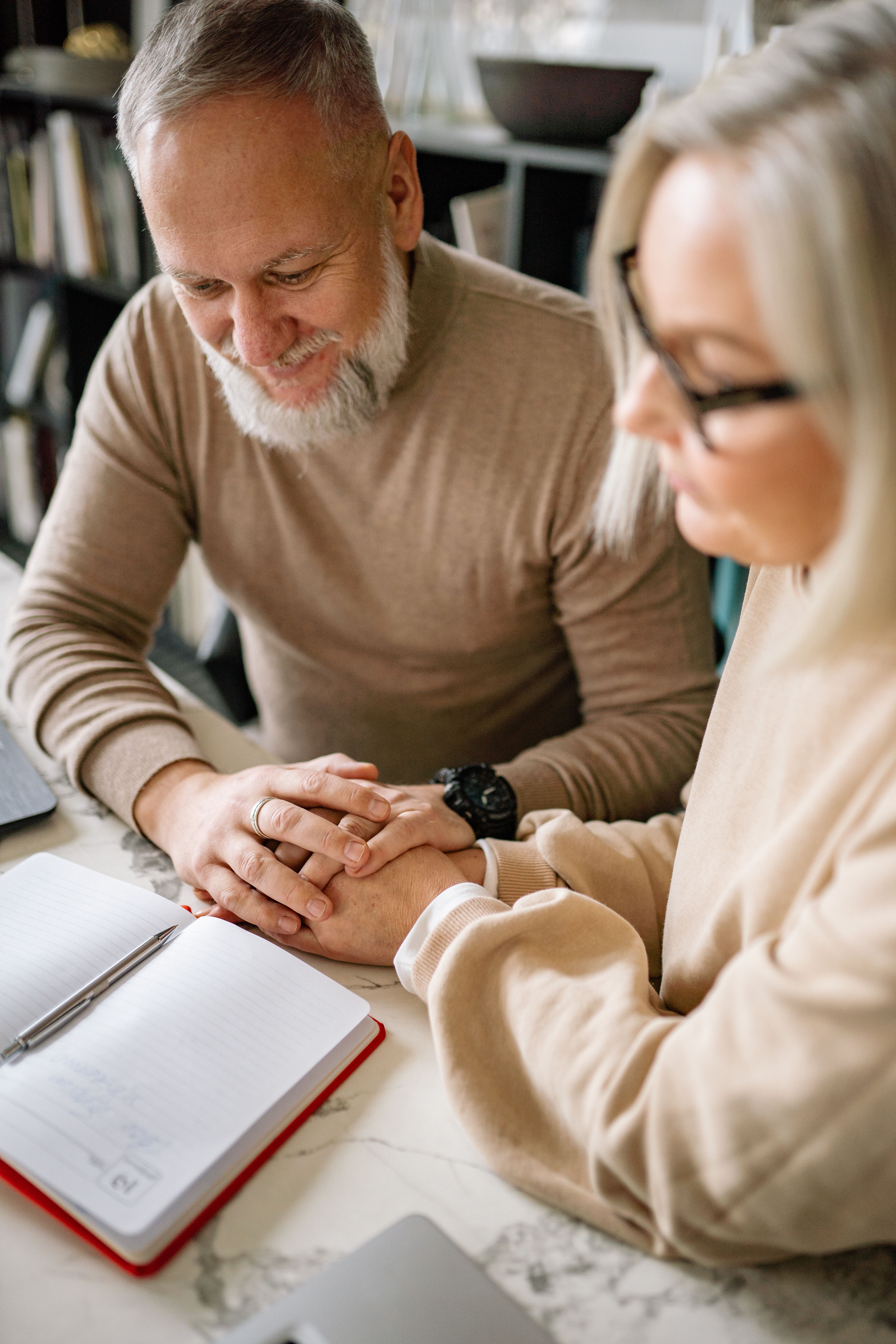 A smiling man and woman sit at a table with their hands clasped over an open notebook and pen, planning together at home.